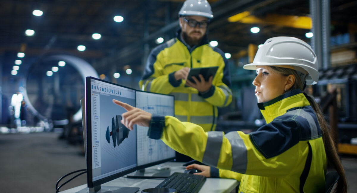 Woman sat at a computer in a ma nufacturing plant, she is qwearing high vis jacket and protective goggles. a male is stood next to her ewearing high vis jacket and goggles